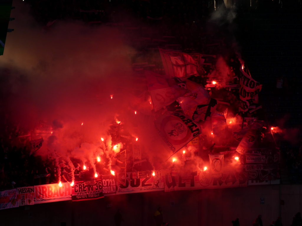 Schwabensturm Fußball-Ultras zünden im Stadion rot leuchtende Pyrotechnik und schwenken Fahnen. Ansonsten ist es dunkel, um sie herum.